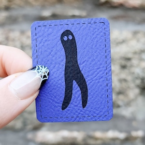 May include: A purple card with a black silhouette of a creature with two eyes and two legs. The card has a stitched border.