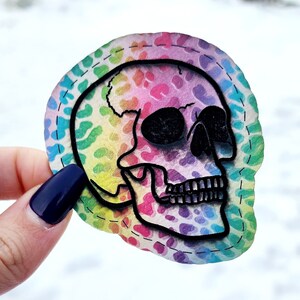 May include: A rainbow-colored skull with black outlines, on a white background. The skull is surrounded by a white border with a stitched pattern.