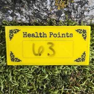 May include: A rectangular yellow sign with black decorative corner accents and the text "Health Points" printed in black. A white rectangular center area features the handwritten number "63" in black ink, positioned against a textured green moss.
