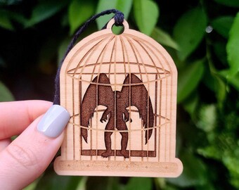 May include: A wooden ornament shaped like a birdcage with a dark silhouette of a winged creature inside. The ornament is hanging from a black string.