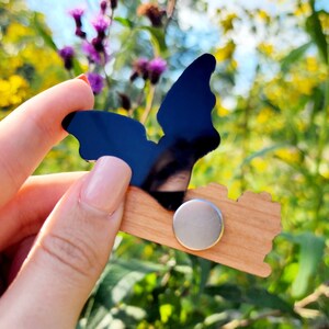 May include: A wooden bat magnet with a black acrylic bat silhouette. The magnet is held in a hand against a background of purple and yellow wildflowers.