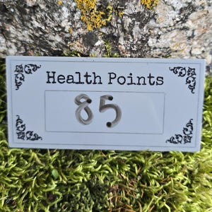 May include: A rectangular white sign with black decorative corner flourishes and the text "Health Points" printed above the number "85" in a central box. The sign features a clean, flat surface with clear, dark lettering and numerals.