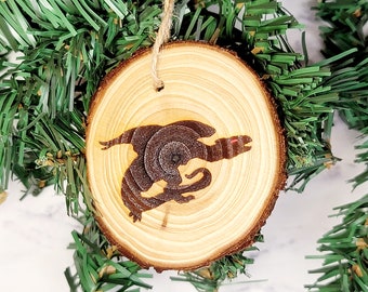 May include: A wooden Christmas ornament with a silhouette of a dragon burned into the surface. The ornament is hanging from a branch of a green artificial Christmas tree.