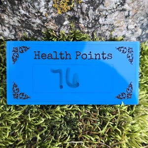 May include: A rectangular bright blue acrylic game tracker with the text Health Points printed in black. The tracker features decorative black corner flourishes and a central rectangular area displaying the handwritten number 76 in black ink.