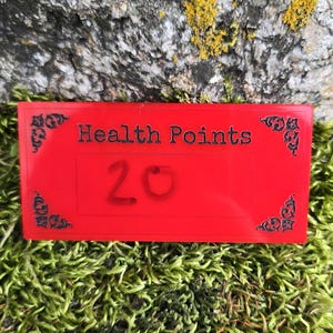 May include: A rectangular red plastic game marker featuring black decorative corner flourishes and the text "Health Points" printed above the number "20" in a black font. The marker has a smooth, flat surface and sharp, clean edges.