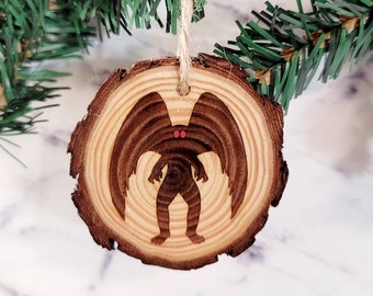 May include: A wooden Christmas ornament shaped like a tree trunk with a silhouette of a creature with wings and red eyes.