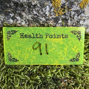 May include: A rectangular, neon yellow acrylic tracker featuring the black text "Health Points" and the number "91" in the center. The corners are decorated with black filigree patterns, and the surface has a smooth, semi-transparent finish.