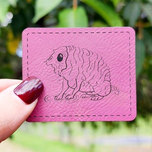 May include: A pink card with a black and white illustration of a creature with a large head and a curly body. The card has a stitched border.