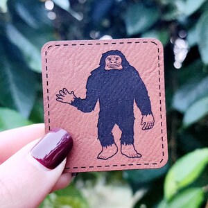 May include: A brown leather patch with a black and white illustration of a smiling Bigfoot waving. The patch has a stitched border.