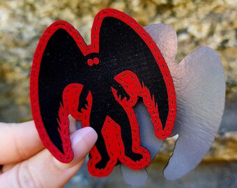 May include: Black and red felt patch with a silhouette of a creature with wings and two red eyes. The patch is attached to a silver backing.