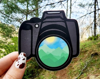 May include: A black and green camera sticker with a viewfinder showing a green and blue mountain landscape with a yellow sun.