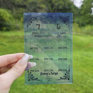 May include: A rectangular, translucent blue spell slot tracker card featuring black printed text and grid boxes. The card displays labels for spell attack bonus, spell save DC, and levels one through nine, with the text Jhotun's Forge at the bottom.