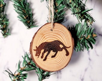 May include: A wooden Christmas ornament with a silhouette of a bull on it. The ornament is hanging from a twine string and is against a white marble background.