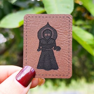 May include: A brown leather patch with a black silhouette of a hooded figure with two eyes. The figure is standing with its arms at its sides.