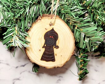 May include: A wooden Christmas bauble featuring a silhouette of a character with a pointed hat and red eyes. The bauble is a natural wood colour with a brown silhouette.