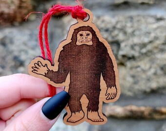 May include: A wooden ornament shaped like a Bigfoot with a red string attached. The Bigfoot is brown and has a toothy grin.