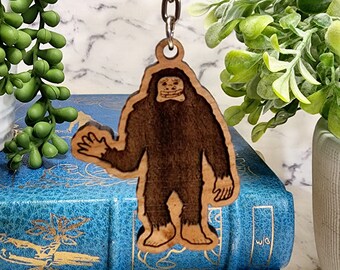 May include: A wooden keychain featuring a cartoon Bigfoot with a friendly expression. The keychain is brown and has a silver chain attached.