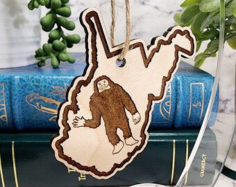 May include: A wooden ornament shaped like the state of West Virginia with a cartoon drawing of a Bigfoot inside. The ornament is hanging from a string.