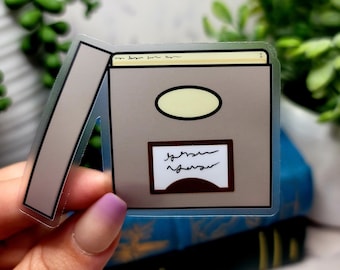 Puede incluir: Una pegatina transparente con un contorno gris de una caja cerrada con una parte superior amarilla. La caja tiene una etiqueta marrón con el texto "german engineer" escrito en negro.