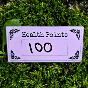 May include: A rectangular light purple sign featuring the black text Health Points and the number 100 centered in a box. The sign is decorated with black ornate corner flourishes on the left and right sides of the top and bottom edges.