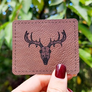 May include: A brown leather coaster with a stitched border features a black silhouette of a deer skull with antlers.