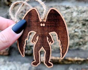 May include: A wooden ornament shaped like a winged creature with large eyes. The ornament is brown and has a rustic finish. It is hanging from a twine string.