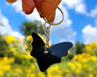 Pode incluir: Um chaveiro de borboleta preto com uma corrente e um anel prateados. O chaveiro é segurado na mão contra um fundo de flores amarelas e céu azul.