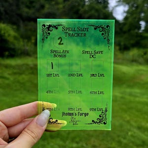 May include: A translucent neon green rectangular spell slot tracker with black engraved text. The board features sections for Spell Slot Tracker, Spell Atk Bonus, Spell Save DC, and levels 1st through 9th, with Jhotun's Forge printed at the bottom.