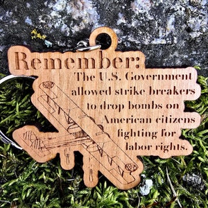 May include: Wooden keychain with a plane design and the text "Remember: The U.S. Government allowed strike breakers to drop bombs on American citizens fighting for labor rights."