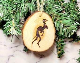 May include: A wooden Christmas ornament with a silhouette of a deer with antlers. The ornament is round and has a natural wood grain. It is hanging from a green garland.