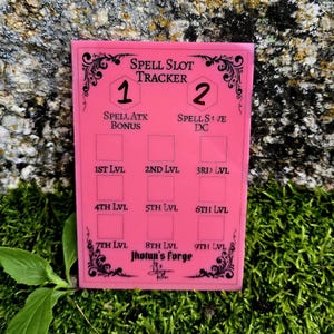 May include: A rectangular pink spell slot tracker board with black text and decorative filigree borders. The board features sections for Spell Slot Tracker, Spell Atk Bonus, Spell Save DC, and levels 1st through 9th, with empty boxes for tracking.