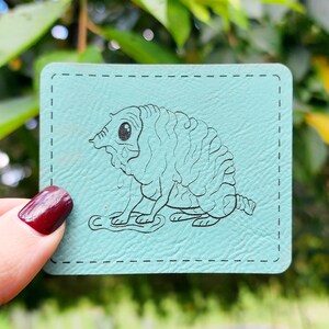 May include: A light blue leather coaster with a black and white line drawing of a creature with a segmented body and large eyes.
