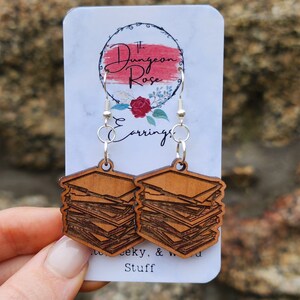 May include: A pair of wooden earrings shaped like stacks of books. The earrings are brown and have a natural wood finish. The earrings are hanging from silver hooks. The earrings are on a white card with the text "The Dungeon Rose Earrings.  Wacky, & Weird Stuff."