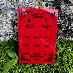 May include: A rectangular red acrylic spell slot tracker featuring black text and decorative filigree. The board includes labels for Spell Slot Tracker, Spell Atk Bonus, Spell Save DC, and levels 1st through 9th, with Jhotun's Forge at the bottom.