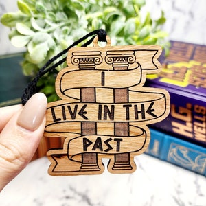 May include: Wooden ornament with a brown wood background and black string. The ornament features two columns with a banner that reads "Live in the Past".