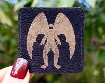 May include: A black leather patch with a gold silhouette of a creature with wings and large eyes. The creature is standing with its wings spread out.