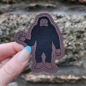 May include: A brown leather patch with a black silhouette of a smiling Bigfoot waving. The patch has a stitched border.