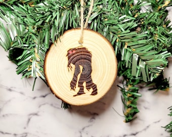 May include: A wooden Christmas ornament featuring a silhouette of a Bigfoot with red eyes. The ornament is round and has a rustic wood grain texture.