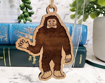May include: Wooden Bigfoot ornament with a smiling face and waving hand. The ornament is made of light-colored wood with a darker brown Bigfoot figure etched into the center. A string is attached for hanging. The ornament is displayed in front of books and greenery.