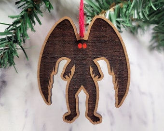 May include: A wooden Christmas ornament shaped like a creature with large wings and red eyes. The ornament is hanging from a red string.