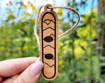May include: A wooden ornament shaped like a snowboard with a mountain scene and three circles engraved on it. The ornament is hanging from a twine string.