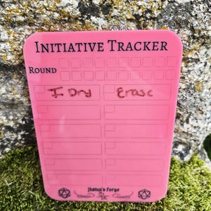 May include: A pink rectangular dry erase initiative tracker board with rounded corners. The board features the text "INITIATIVE TRACKER" at the top, a "ROUND" counter, and multiple blank rows for writing. The text "Jotun's Forge" is printed at the bottom.