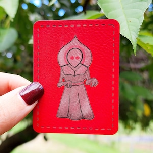 May include: A red card with a white stitched border. The card features a drawing of a person wearing a hooded robe with a red face and two eyes. The person is holding their hands out in front of them.