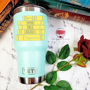 May include: A turquoise Yeti tumbler with a stainless steel lid and base. The tumbler has a graphic of stacked books with the text "Support Public Libraries" in black lettering.