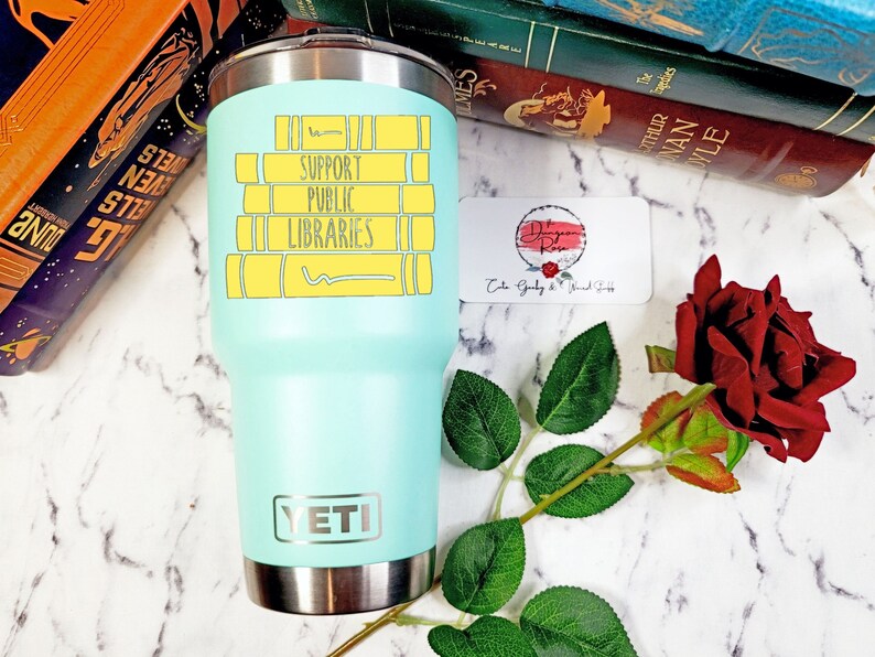 May include: A turquoise Yeti tumbler with a stainless steel lid and base. The tumbler has a graphic of stacked books with the text "Support Public Libraries" in black lettering.