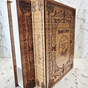 Puede incluir: Una caja de madera con forma de libro, con una cubierta frontal de madera clara y un lomo de madera oscura. La cubierta presenta bordes ornamentados grabados con l&aacute;ser y el texto Jhotun's Forge by The Dungeon Rose. El lomo muestra el texto The Dungeon Rose.