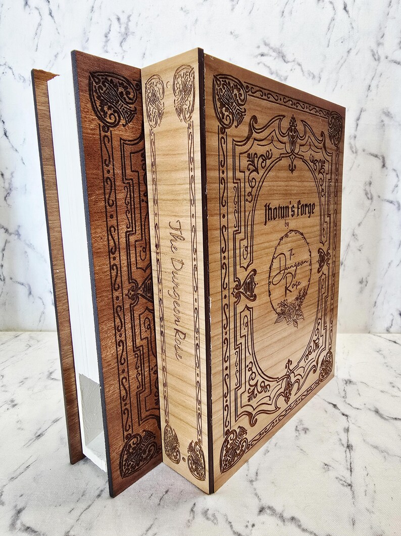 Puede incluir: Una caja de madera con forma de libro, con una cubierta frontal de madera clara y un lomo de madera oscura. La cubierta presenta bordes ornamentados grabados con l&aacute;ser y el texto Jhotun's Forge by The Dungeon Rose. El lomo muestra el texto The Dungeon Rose.