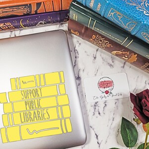 May include: A silver laptop with a yellow sticker that says "Support Public Libraries". The sticker is shaped like a stack of books. The laptop is on a white marble surface with a stack of books and a red rose.
