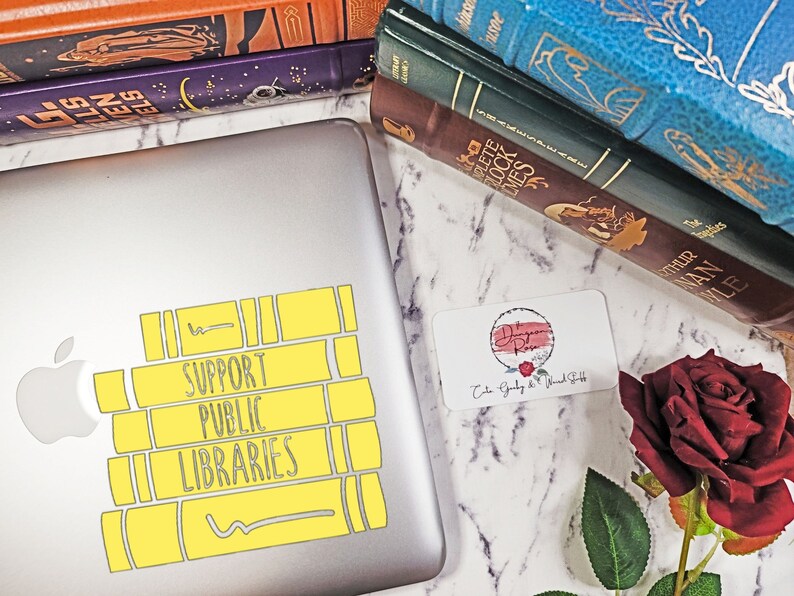 May include: A silver laptop with a yellow sticker that says "Support Public Libraries". The sticker is shaped like a stack of books. The laptop is on a white marble surface with a stack of books and a red rose.