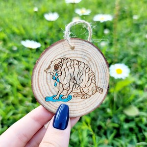 May include: A wooden ornament shaped like a tree trunk with a hand-painted illustration of a cartoon animal with blue eyes and a tear. The animal is sitting on a blue puddle.
